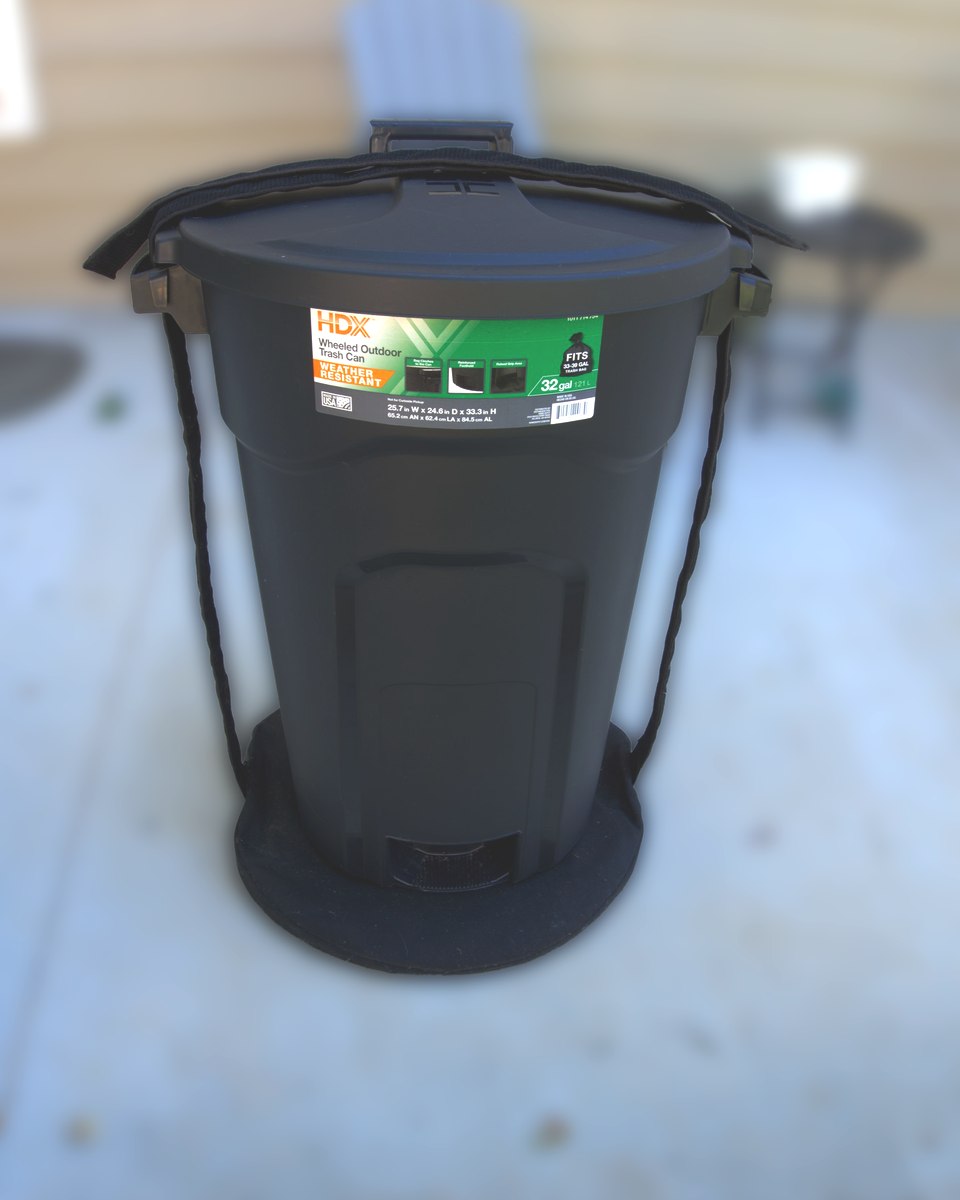 Front view of a full-size trash can held down on the round black CanClamp base, straps running up both sides and over the lid, background softly blurred